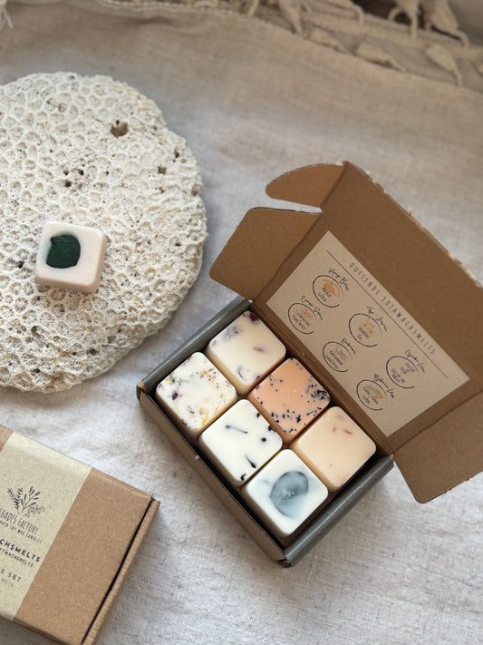 A box of scented wax melts from Shades Factory, with individual melts visible, hand-poured with dried flowers on top, displayed next to a white textured object.