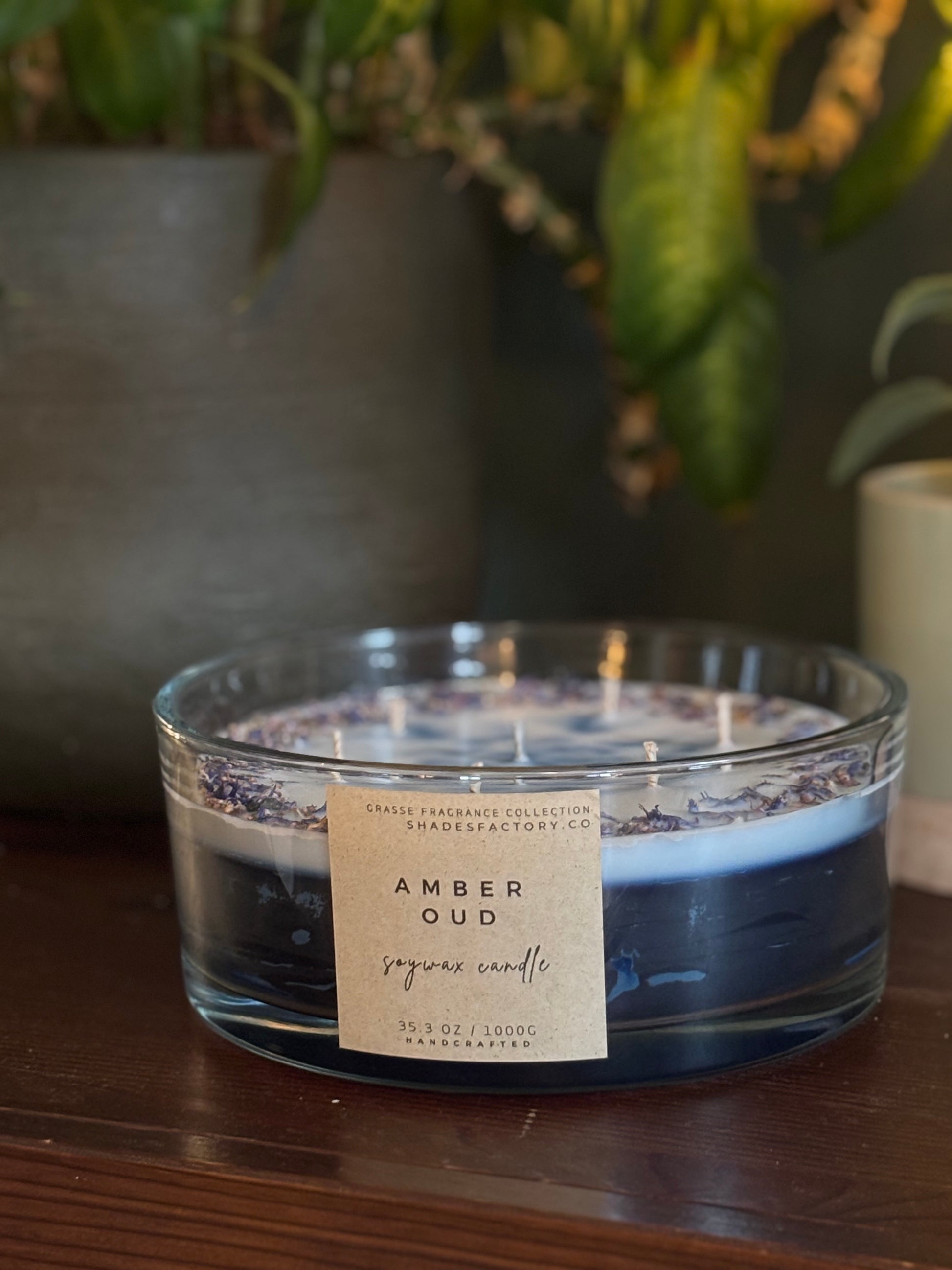 Candle labeled 'Amber Oud' on a wooden surface with a blurred plant in the background