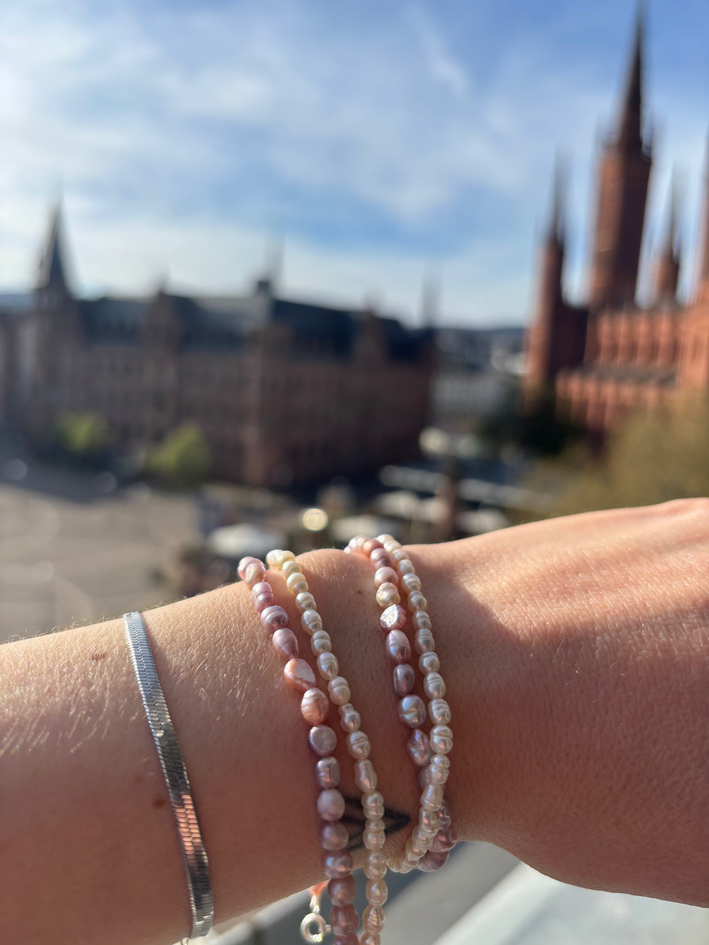 Close-up of a wrist wearing multiple pearl bracelets with a blurred background of a cityscape.