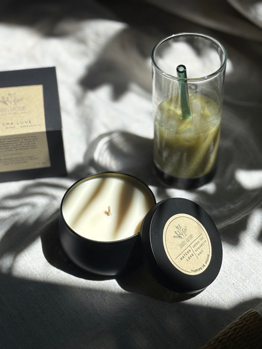 Matcha candle in a black container with a label, next to a glass of iced drink on a textured surface.